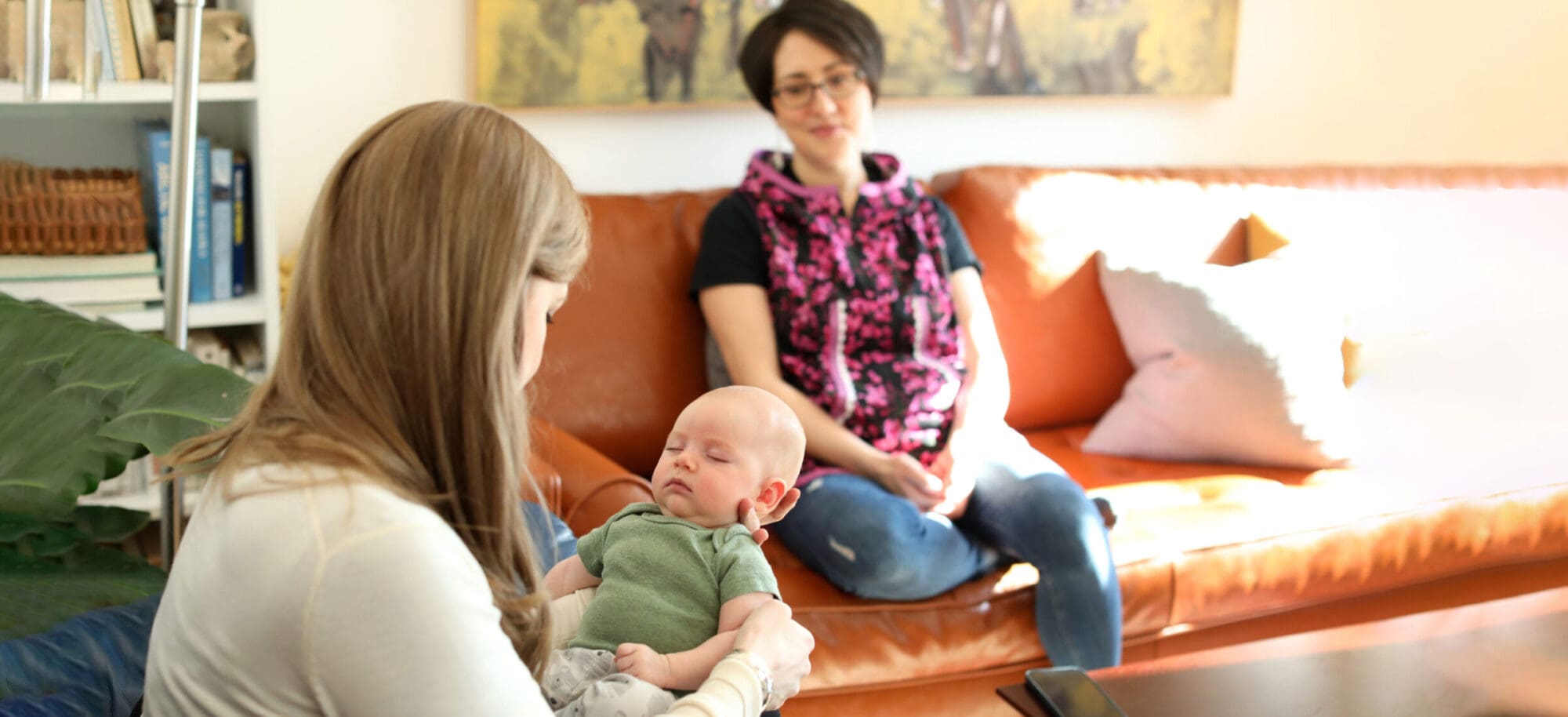 Image of Penny Gage on a couch holding her child, Loretta, in a beautiful sunlit living room, with Lena sitting on another couch adjacent to the pair looking gently at the baby.
