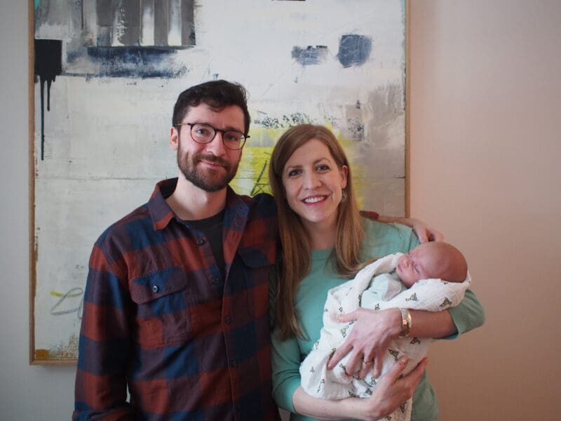 Zach and Penny standing together in front of a whiteboard, smiling, while Penny holds their baby, Loretta.