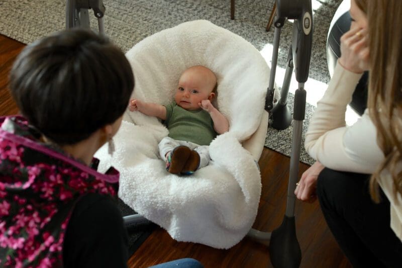Two women (Lena and Penny) looking at a newly born baby, stretching in a plushy white crib.